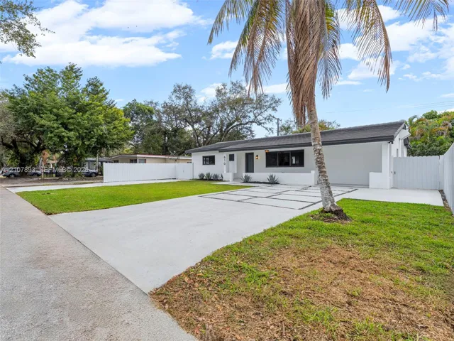 Single-story exterior featuring a painted stucco finish, attached carport, grid-patterned concrete driveway, white perimeter fencing, and established palm trees