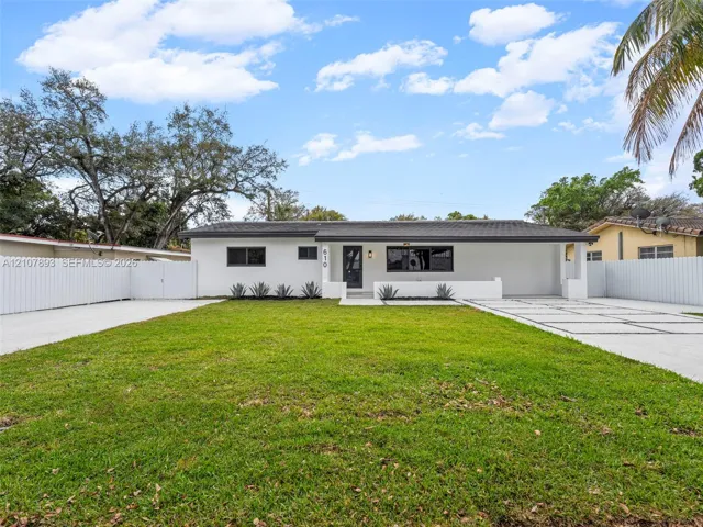 Single-story exterior featuring white stucco siding, a low-slope roof, expansive front lawn, concrete paver driveway with integrated turf, and full-perimeter privacy fencing