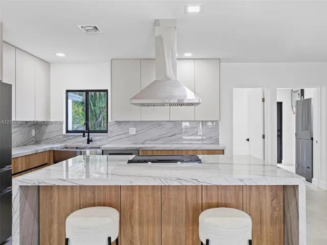 Kitchen featuring a waterfall-edge stone island with a cooktop, wood-finish cabinetry, stone slab backsplash, stainless steel chimney range hood, and matte black faucet