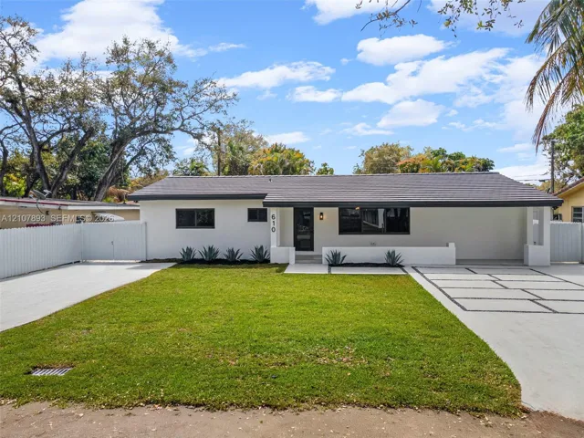 Single-story exterior featuring white stucco finish, dark-toned roof, paved geometric driveway, white perimeter fencing, and manicured lawn