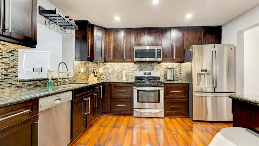 Kitchen featuring dark wood cabinetry, stainless steel appliances, multi-tonal stone tile backsplash, stone countertops, and wood-finish flooring