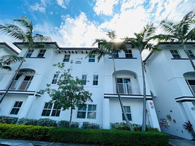 Multi-story exterior featuring white stucco facade, arched architectural detailing, private balconies with vertical railings, and mature palm tree landscaping