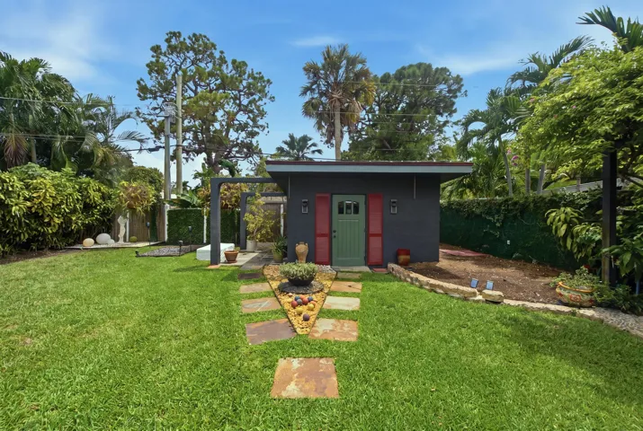 Detached structure featuring a masonry exterior, decorative red shutters, a green paneled entry door, a stone-paved walkway, and mature landscaping