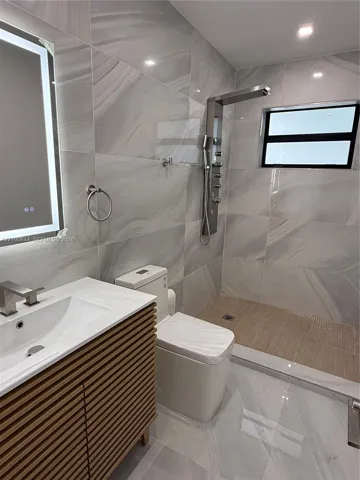 Bathroom featuring large-format wall tiles, a wood-slat vanity with a white countertop and rectangular sink, a walk-in shower with a multi-head stainless steel panel, and an integrated LED-lit vanity mirror