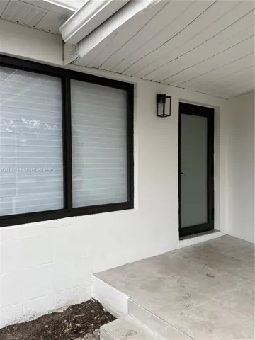 Exterior entryway featuring white painted concrete block construction, large window with dark frame, frosted glass door, tile porch surface, and wall-mounted black metal lantern fixture