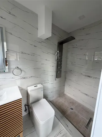 Bathroom featuring floor-to-ceiling veined stone-finish tile, walk-in shower with a stainless steel multi-head spray system, slatted-wood finish vanity, and integrated-tank toilet