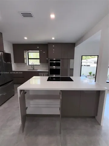 Kitchen featuring a waterfall-edge center island with a cooktop, slab-front cabinetry, stainless steel wall-mounted oven and microwave, recessed lighting, and large-format tile flooring