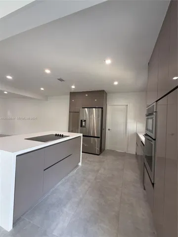 Kitchen featuring gloss-finish cabinetry, an island with a waterfall-edge countertop and integrated cooktop, large-format tile flooring, and stainless steel built-in appliances including a wall oven and refrigerator