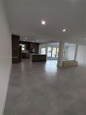 Open-concept interior featuring large-format tile flooring, recessed ceiling lighting, a central kitchen island with waterfall-edge countertop, and floor-to-ceiling windows with black framing