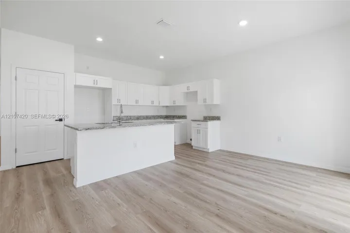 Kitchen featuring white shaker-style cabinetry, stone-finish countertops, an island with an integrated stainless steel faucet, recessed lighting, and wood-finish flooring