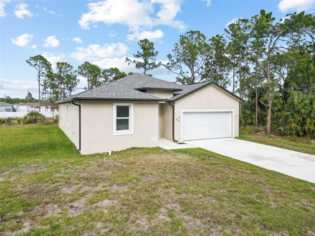 Single-story stucco exterior featuring a hip roof, two-car garage, concrete driveway, and surrounding mature trees