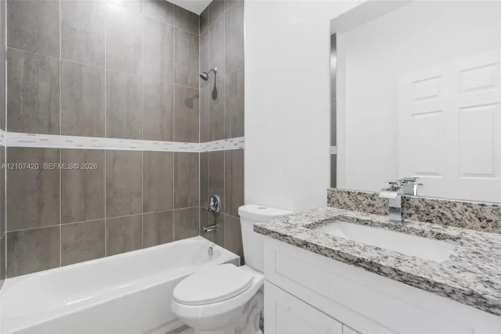 Bathroom featuring a vanity with a stone countertop, an integrated rectangular sink with a modern chrome faucet, a white toilet, and a bathtub with a shower head and full-height wall tile surround with decorative accent tiling
