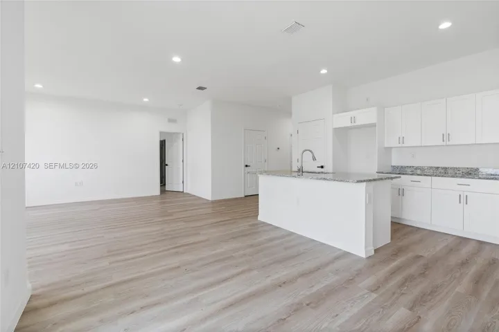 Open-concept kitchen featuring white shaker-style cabinetry, stone-surface countertops, a center island with a stainless steel pull-down faucet, and wood-finish flooring