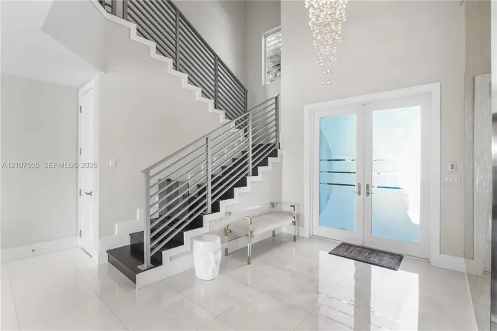 Foyer featuring high ceilings, large-format tile flooring, a double-door entry with frosted glass detailing, a contemporary tiered crystal chandelier, and an open-riser staircase with horizontal metal railing