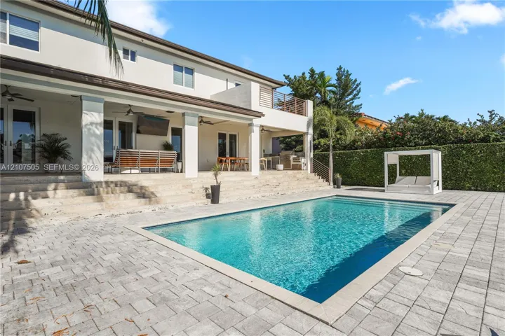Covered rear patio with stone-finish steps, rectangular swimming pool, concrete paver decking, and upper-level balcony with horizontal railing