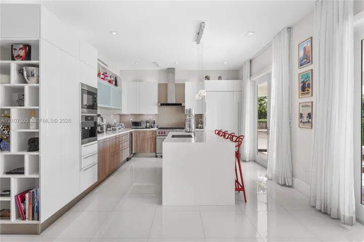 Kitchen featuring custom flat-panel cabinetry, a central island with a solid-surface countertop, built-in stainless steel appliances, a stainless steel range hood, and large-format porcelain tile flooring