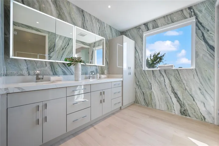 Dual-sink vanity with high-gloss cabinetry, stone-surfaced countertops, full-wall patterned stone cladding, and triple-panel mirrored medicine cabinets, complemented by wood-finish flooring