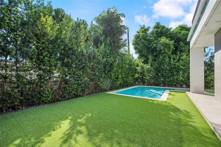 Private backyard featuring an in-ground swimming pool, synthetic turf lawn, stone paver decking, and mature perimeter hedging
