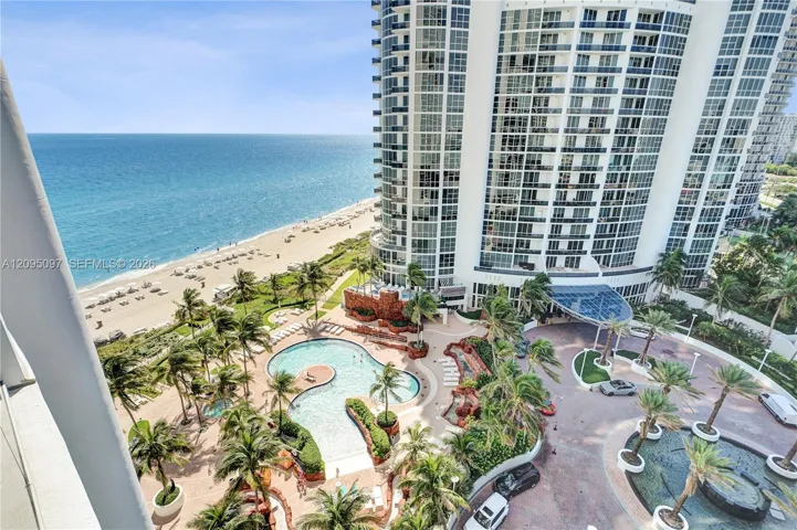 High-rise architectural exterior featuring glass-paned facades, a landscaped courtyard with a curvilinear swimming pool, mature palm trees, a circular driveway, and direct oceanfront positioning