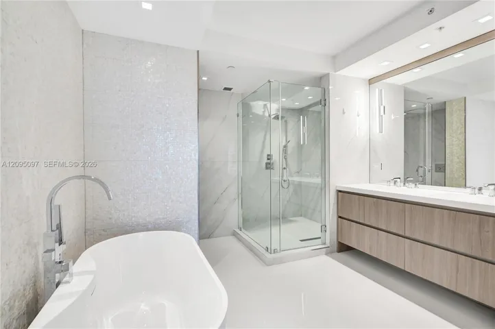 Freestanding soaking tub with floor-mounted faucet, glass-enclosed shower, wall-to-wall vanity with double sinks, and recessed lighting