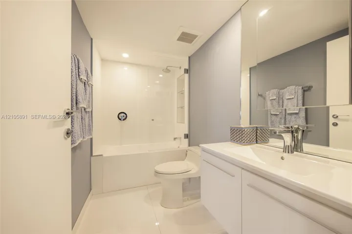 Bathroom featuring a white vanity with an integrated sink, a large wall-mounted mirror, a tub-shower combination with a glass panel, and polished tile flooring