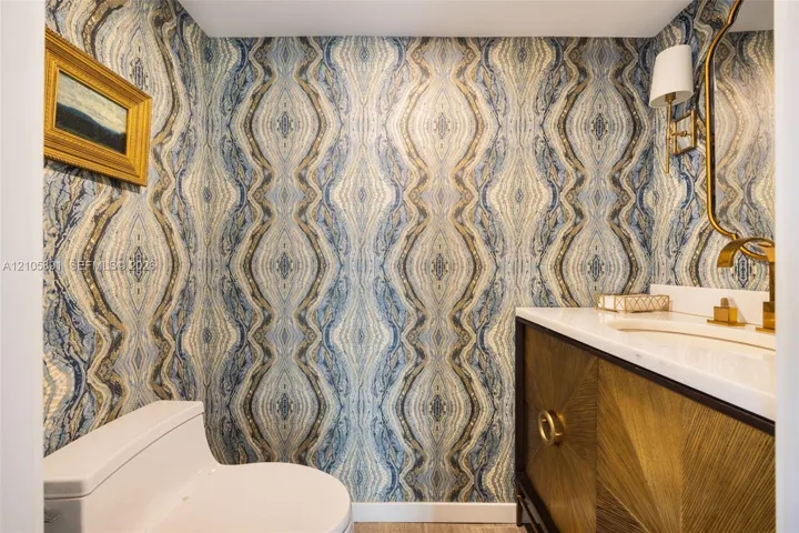 Powder room featuring patterned wallpaper, a wood-finish vanity with sunburst detailing, a stone-composite countertop, an integrated sink, and brass-finish fixtures