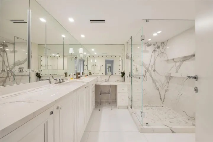 Dual-sink vanity with quartz countertop, expansive wall-to-wall mirrors, integrated makeup vanity station, and glass-enclosed walk-in shower featuring floor-to-ceiling stone wall cladding