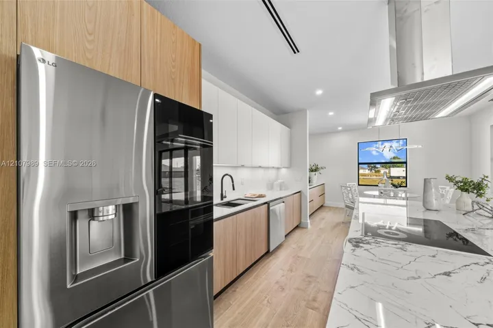 Kitchen featuring wood-finish cabinetry, white upper cabinets, stone-surfaced island with waterfall edge, stainless steel refrigerator, and recessed lighting
