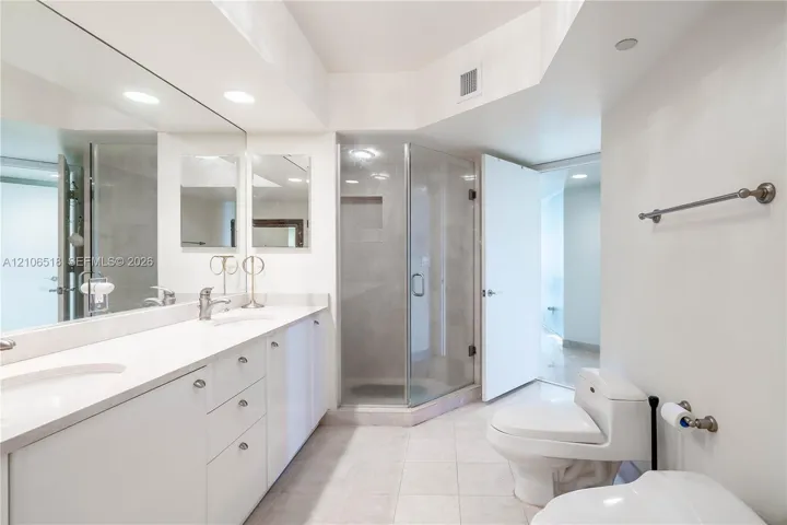 Double vanity with white cabinetry and solid-surface countertop, full-width wall mirror, glass-enclosed corner shower with tiled interior, and ceramic tile flooring