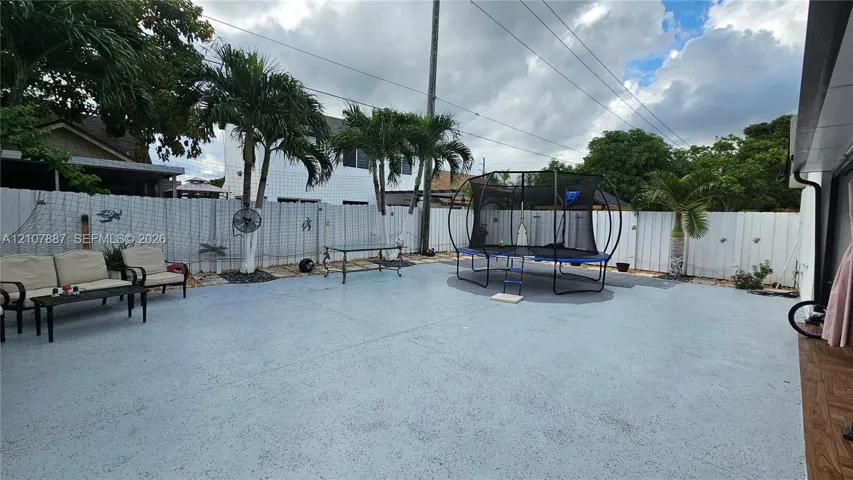 Spacious concrete patio featuring a white vinyl privacy fence, mature palm trees, and exterior wall-mounted lighting