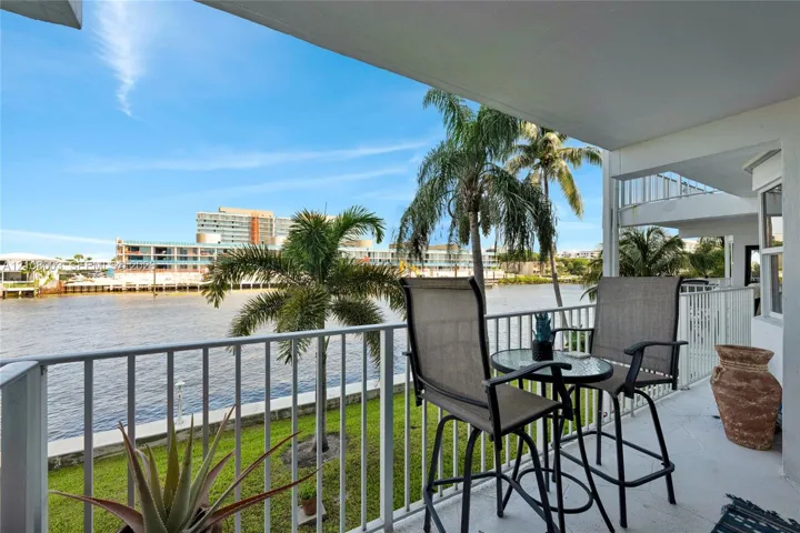 Covered balcony featuring white vertical railing and concrete flooring, overlooking the waterfront with mature palm trees and manicured lawn