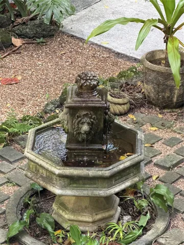 Hexagonal stone water fountain featuring lion head sculptural details, integrated into a landscape setting with paver accents and gravel ground cover