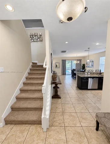 Carpeted staircase with white painted balustrade, tile flooring, kitchen featuring dark cabinetry and stainless steel appliances, and ceiling-mounted light fixtures