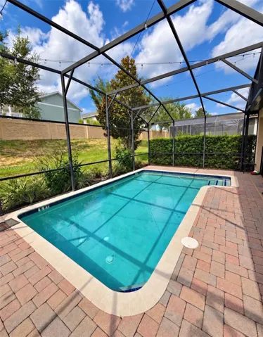 Screen-enclosed swimming pool featuring a paver deck, perimeter hedge, and string lighting