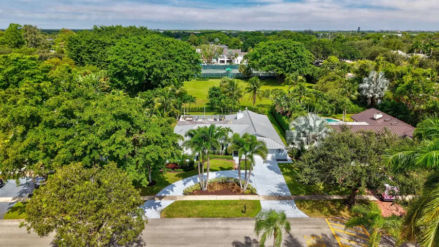 Aerial view featuring a metal roofing system, paved driveway, landscaped perimeter, and mature canopy trees