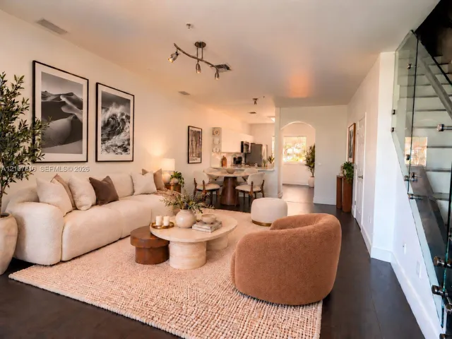 Open-concept interior featuring dark-tone wood-finish flooring, a contemporary multi-bulb ceiling fixture, a glass-panel staircase railing, and a kitchen area with white cabinetry and an arched entryway