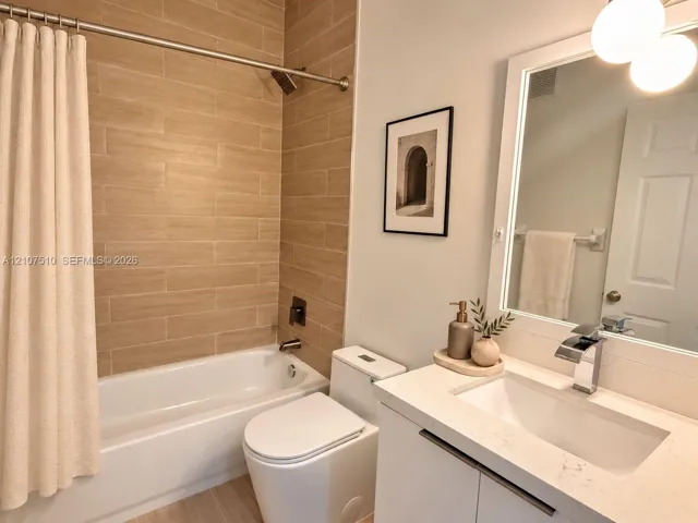 Bathroom featuring a light-toned, plank-tile shower surround, a white vanity with a solid-surface countertop, an integrated rectangular sink, a polished metal waterfall faucet, and a wall-mounted light fixture