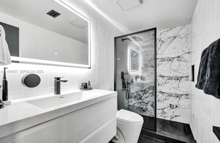 Modern bathroom featuring a floating vanity with an integrated rectangular sink, wall-mounted matte black faucet, illuminated vanity mirror, and a walk-in shower with stone-patterned wall tile and black metal framing