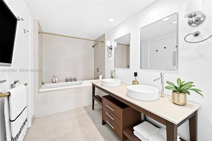 Dual-vanity bathroom featuring a wood-frame vanity with a stone countertop, two vessel sinks, wall-mounted faucets, and a deep soaking tub with tile surround -