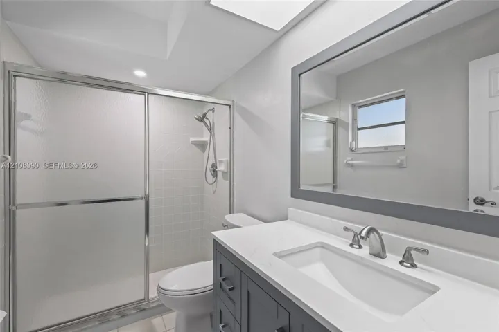 Bathroom featuring a charcoal-toned vanity with a white countertop and integrated sink, a large framed mirror, a walk-in shower with a sliding glass enclosure, and a skylight