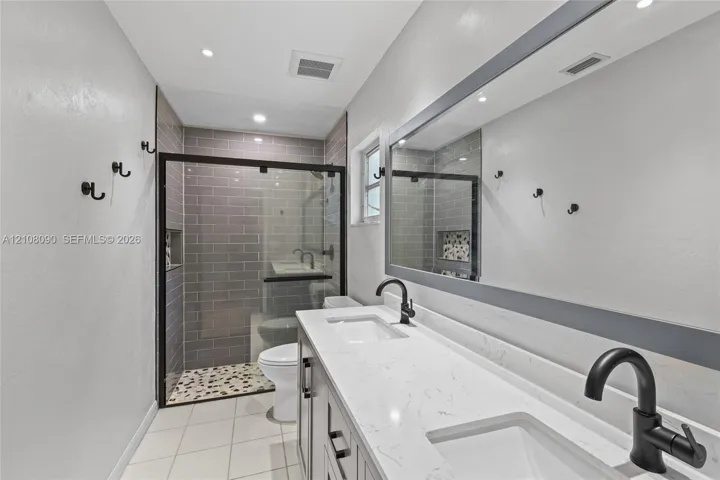 Double vanity featuring white stone-finish countertops, undermount sinks, matte black fixtures, full-width mirror, and a glass-enclosed shower with grey subway tile surround and pebble tile flooring