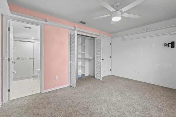 Bedroom featuring carpeted flooring, built-in closet shelving, a white ceiling fan with integrated lighting, and access to a bathroom with a glass-enclosed shower stall
