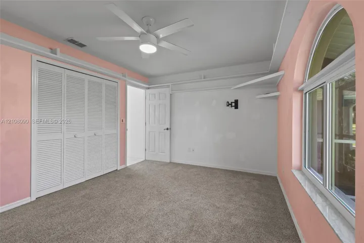 Wall-to-wall carpeting, white louvered-style closet doors, arched window with stone-finish sill, ceiling fan with integrated lighting, and built-in wall shelving