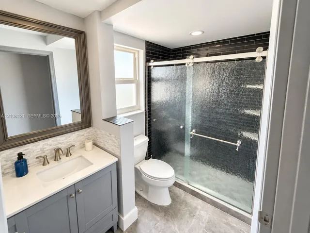 Bathroom featuring a gray vanity with a stone countertop, an integrated rectangular sink, textured tile backsplash, a walk-in shower with black subway tile surround and sliding glass doors, and recessed lighting