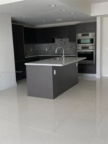 Kitchen featuring dark cabinetry, quartz countertops, a central island with a stainless steel faucet, stacked built-in stainless steel ovens, and large-format tile flooring