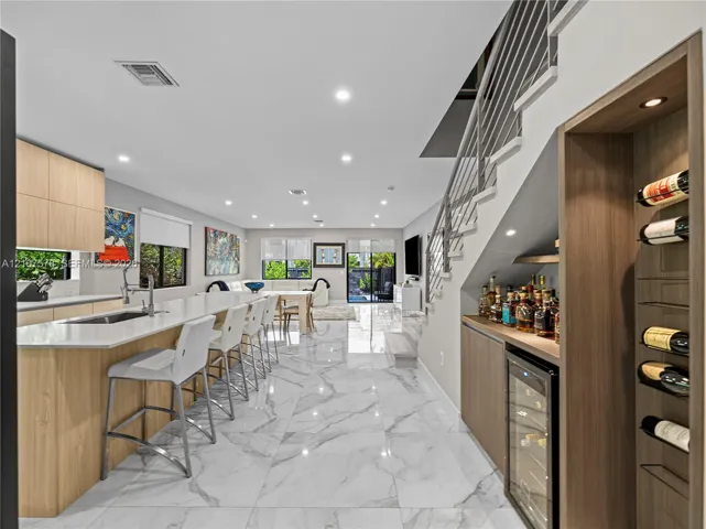 Open-concept kitchen featuring light wood-finish cabinetry, stone-surface countertops, large-format stone-finish flooring, a built-in wine cooler with integrated wine rack, and a staircase with metal railing