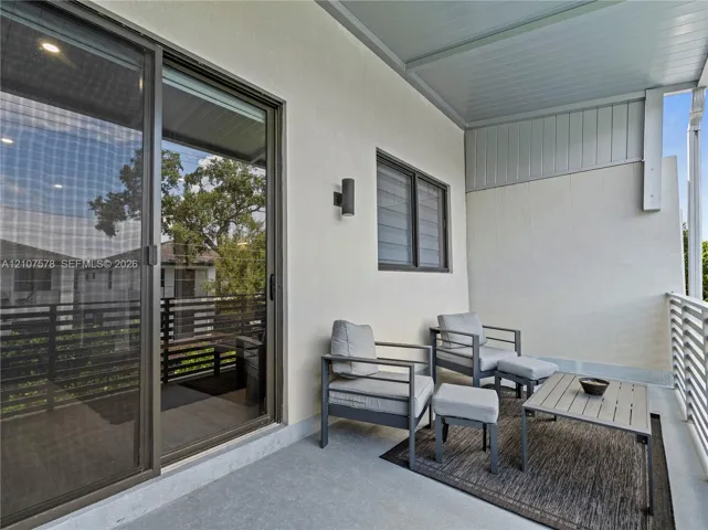 Covered balcony featuring sliding glass doors, horizontal slat railings, neutral stucco exterior wall, and wall-mounted exterior lighting