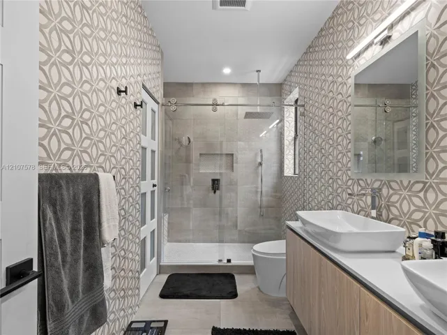 Floating wood-finish vanity with dual vessel sinks, walk-in shower with glass enclosure and rain-style showerhead, patterned wall tile, and built-in recessed shower niche