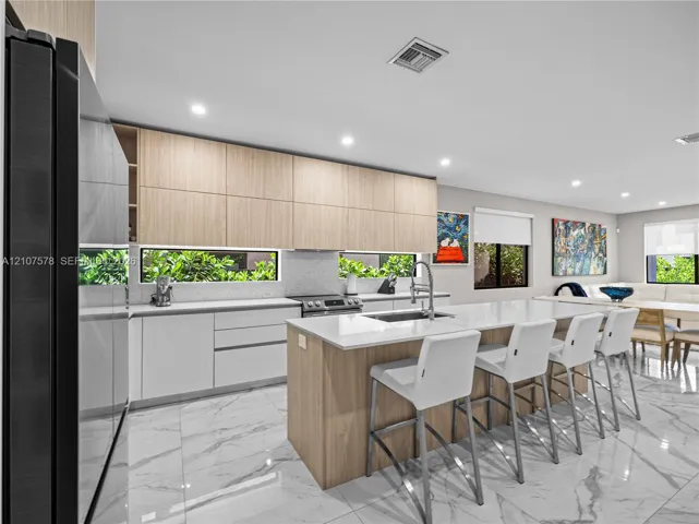 Kitchen featuring flat-panel wood and white cabinetry, stone-finish countertops, a central island with an integrated sink, and light-toned tile flooring