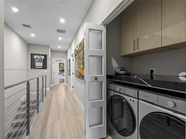 Laundry closet featuring light-toned cabinetry, high-gloss solid surface countertop, and recessed ceiling lighting, accompanied by a hallway with wood-finish flooring and a metal cable railing system